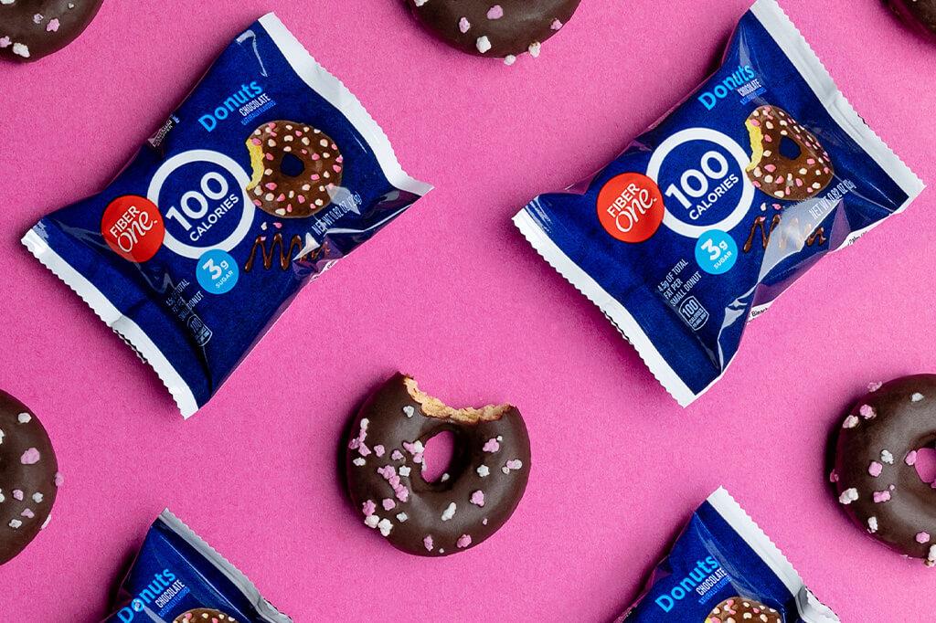 Packaged chocolate frosted donuts with sprinkles and a bitten donut, displayed on a pink background.