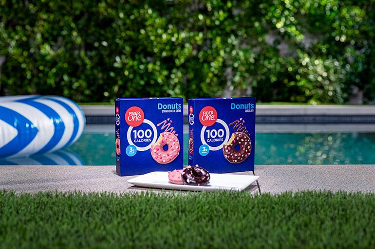 Two blue boxes of FiberOne Strawberry & Creme and Chocolate Donuts, next to a pool with green grass