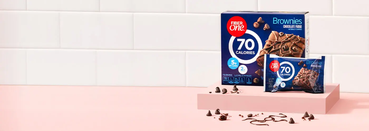 A box and a single packaged bar of Fiber One Chocolate Fudge Brownies, raised on a pink platform on a kitchen countertop