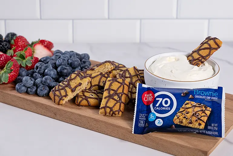 A wooden cutting board with a bowl of Cheesecake Dip, a bar of Fiber One Chocolate Chip Brownie and assorted berries