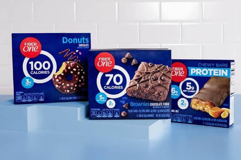 Three boxes of Fiber One products are displayed: chocolate donuts (100 calories), chocolate fudge brownies (70 calories), and protein chewy bars with caramel and nuts (5g protein per serving), against a blue and white background.